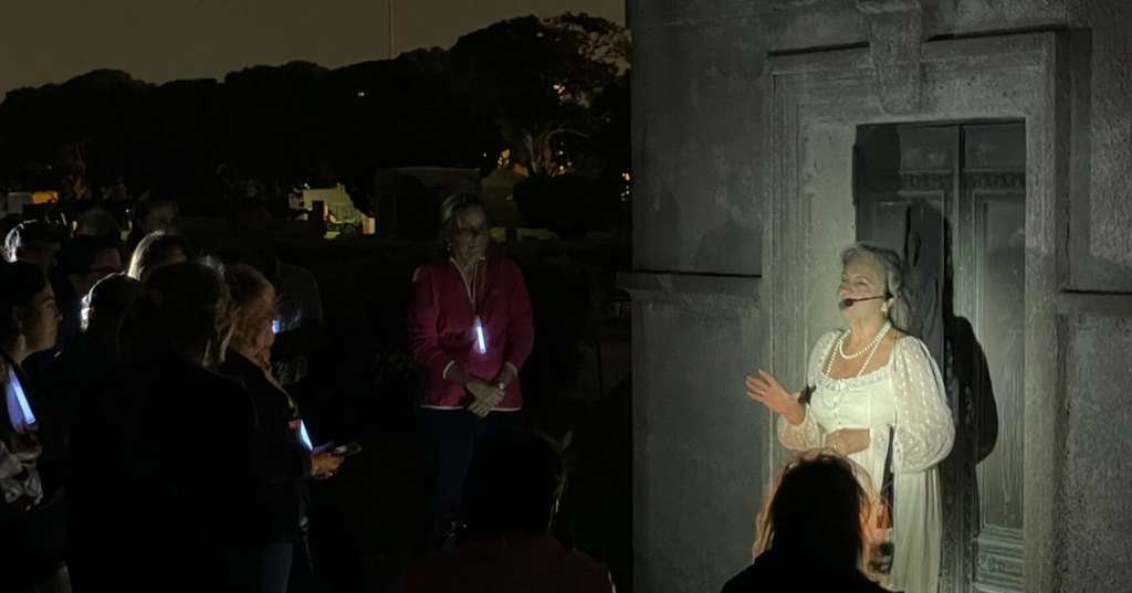 A group of people in a cemetery listening to a woman in period dress who is speaking.
