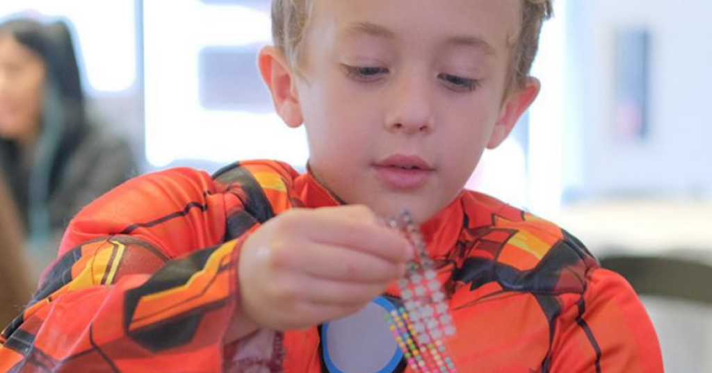 A young boy wearing a colorful superhero costume is focused on creating something with colorful gems.