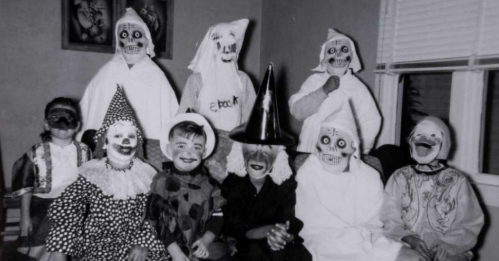 A group of children and adults dressed in various Halloween costumes, including clowns, witches, and ghostly figures, posing together in a room.