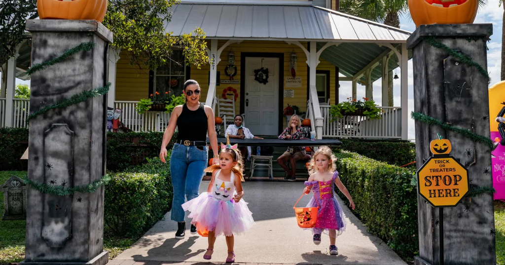 Two children in costumes are walking from a house in Yesteryear Village while an adult accompanies them. The entrance is decorated for Halloween with pumpkins and signs.